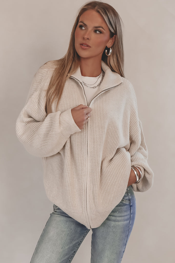 Woman wearing a beige zip-up sweater and light blue jeans against a plain background