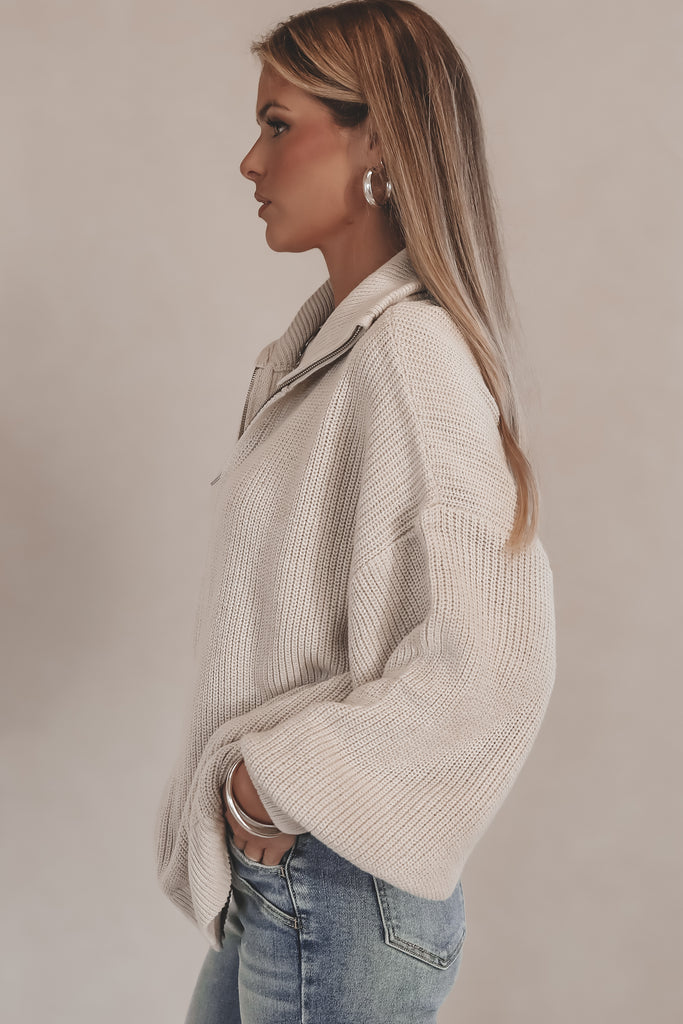 Woman wearing a beige sweater and blue jeans against a plain background