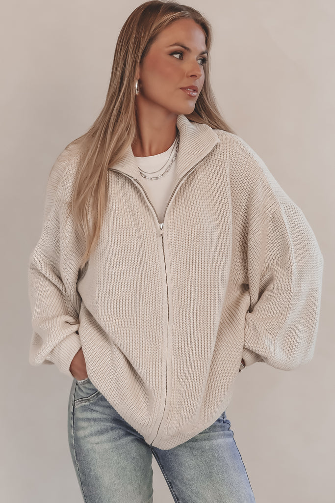 Woman wearing a beige zip-up sweater and jeans against a plain background