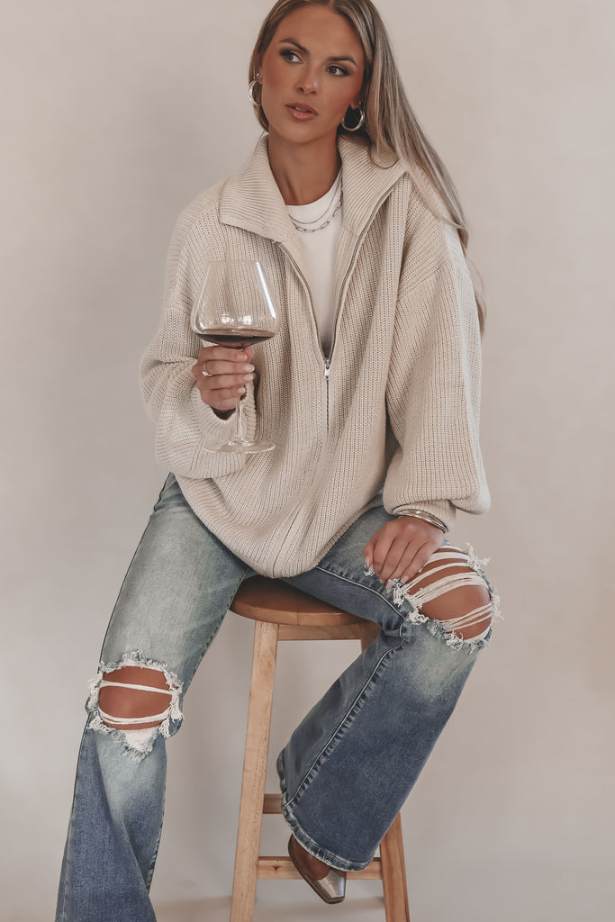 Woman sitting on a stool holding a glass of red wine, wearing a beige jacket and ripped jeans.