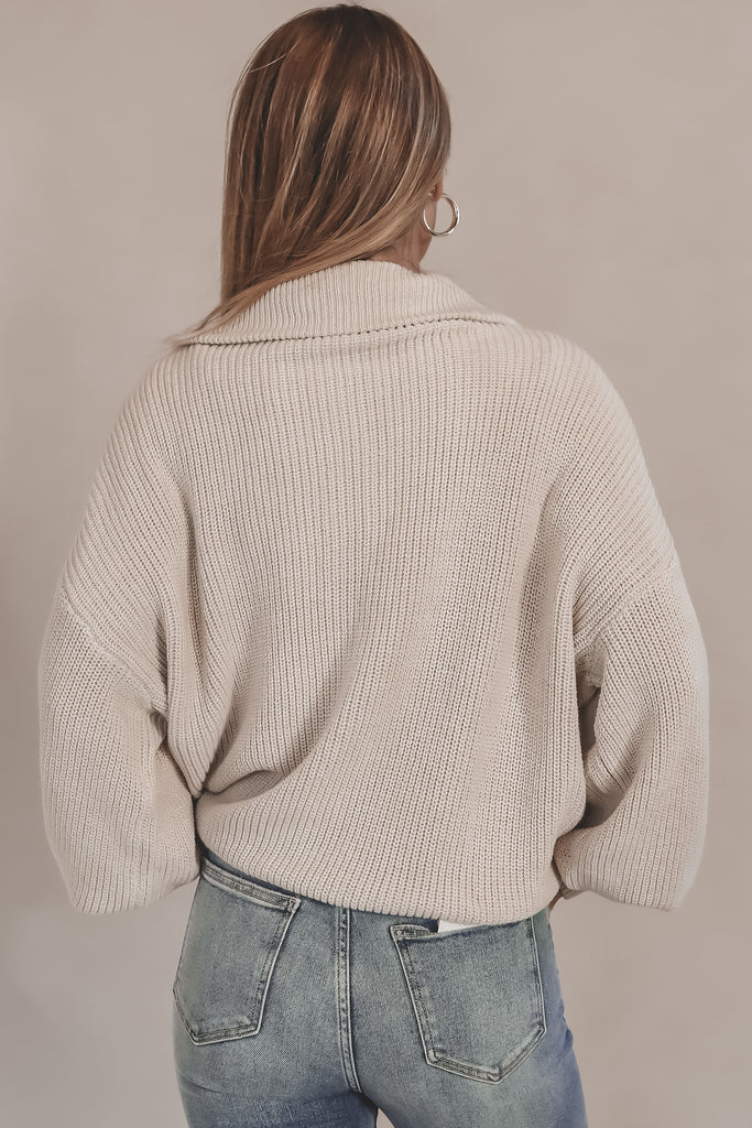 Person wearing a beige knitted sweater and blue jeans against a plain background
