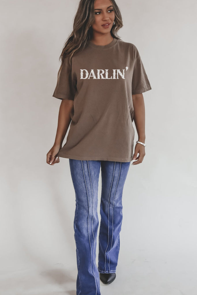 Woman wearing a brown t-shirt with 'DARLIN'' text and blue jeans on a plain background