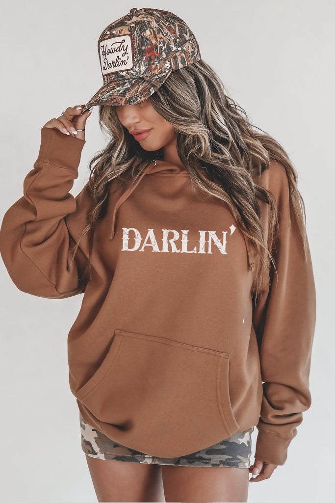 Person wearing a brown 'Darlin' hoodie and camouflage cap on a plain background