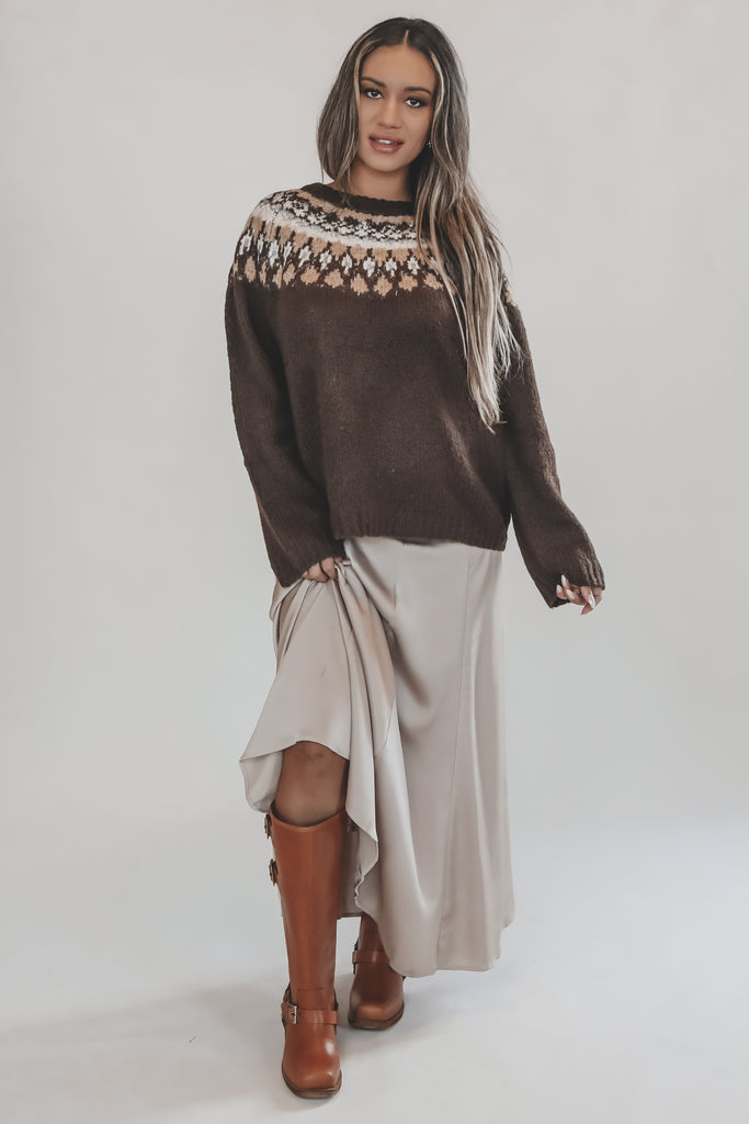 Woman wearing a brown sweater with a patterned neckline, beige skirt, and brown boots on a white background