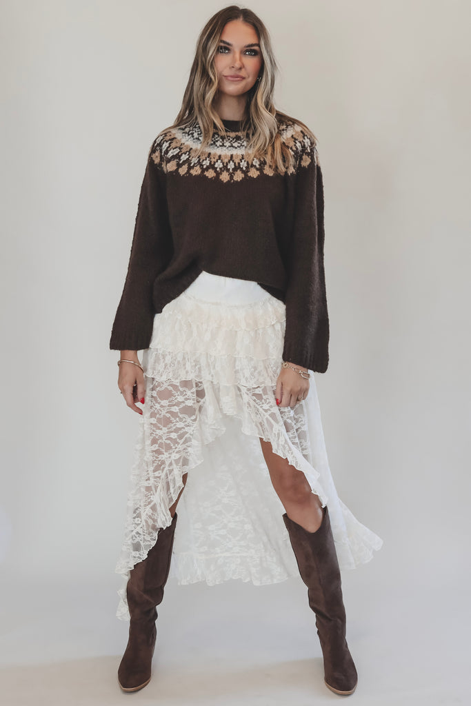 Woman wearing a brown sweater with a patterned neckline, white lace skirt, and brown knee-high boots on a plain background.