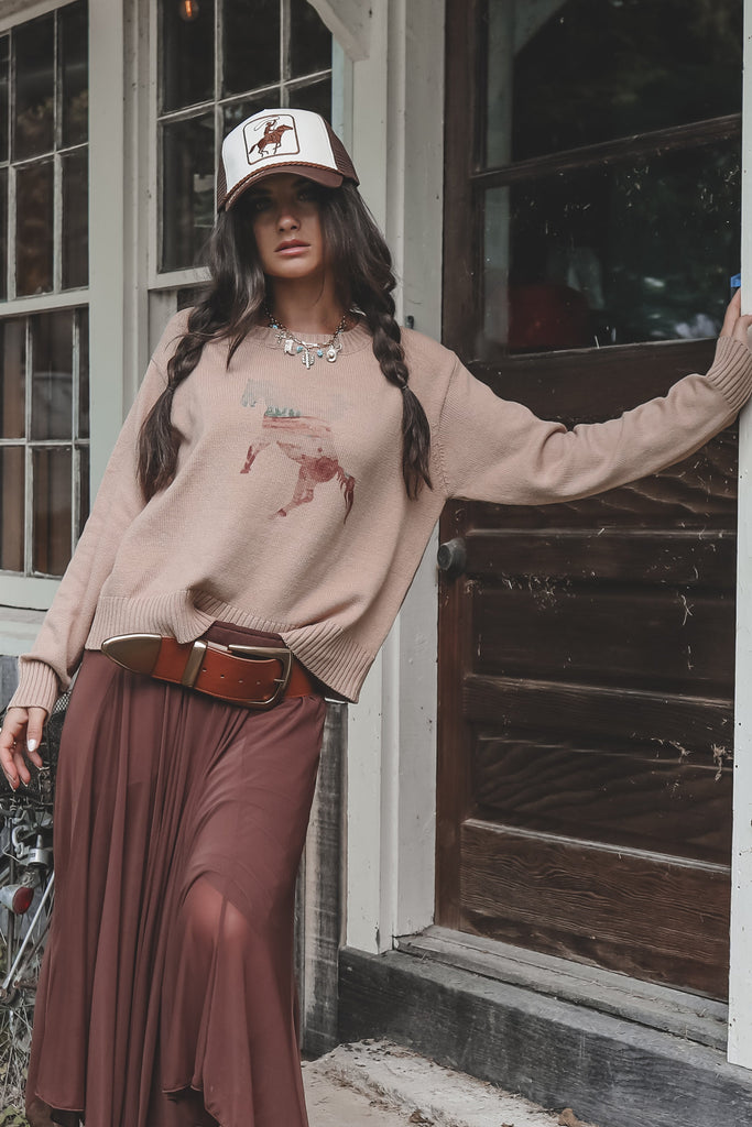 Woman standing on a porch wearing a beige sweater and brown skirt.
