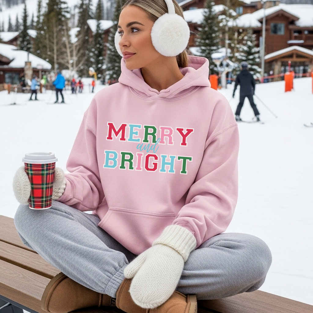Person wearing a pink hoodie with 'Merry and Bright' text, sitting on a bench in a snowy outdoor setting.