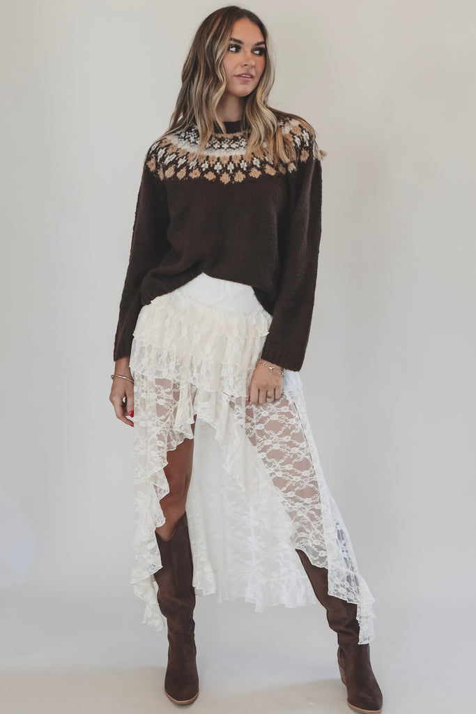 Woman wearing a dark sweater with a patterned neckline, white lace skirt, and brown boots on a plain background