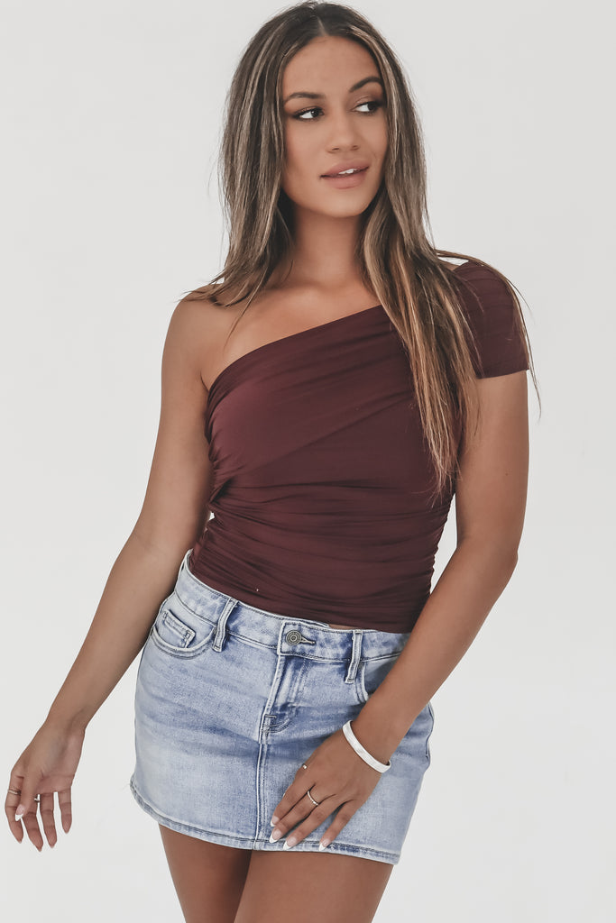 Woman wearing a one-shoulder burgundy top and denim skirt on a white background