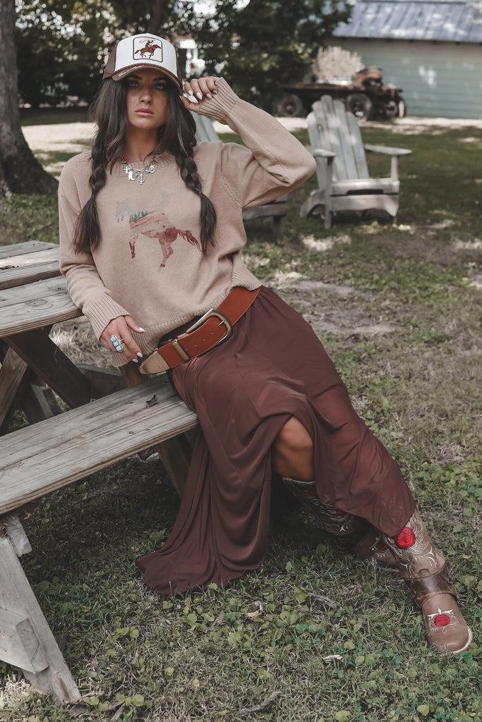 Woman sitting on a wooden bench outdoors wearing a beige sweater, brown skirt, and cowboy boots.
