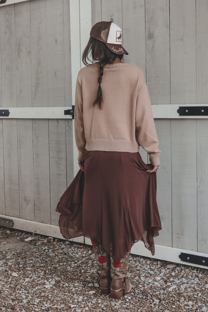 Person wearing a beige sweater and brown skirt standing against a wooden wall.