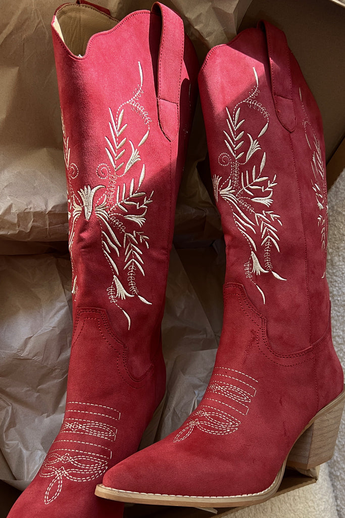 Red cowboy boots with white embroidery in a cardboard box on a textured surface.