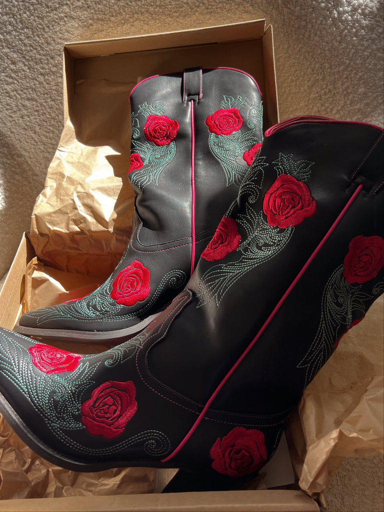 Black cowboy boots with red rose embroidery in a box on a textured surface.