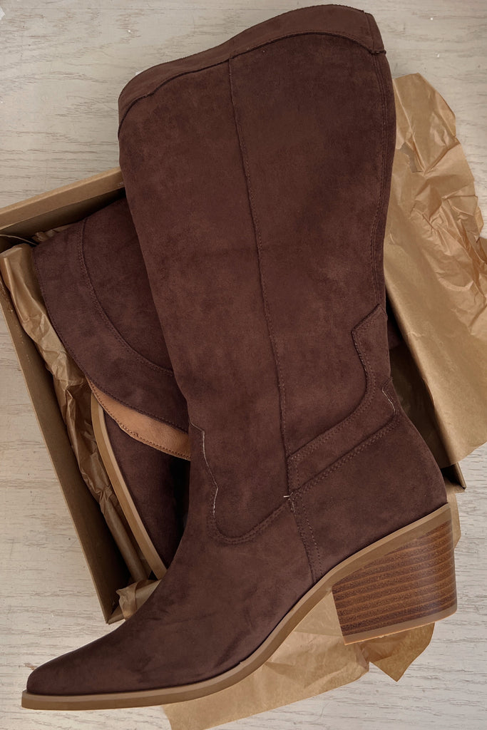 Brown suede knee-high boots in a cardboard box on a wooden floor.