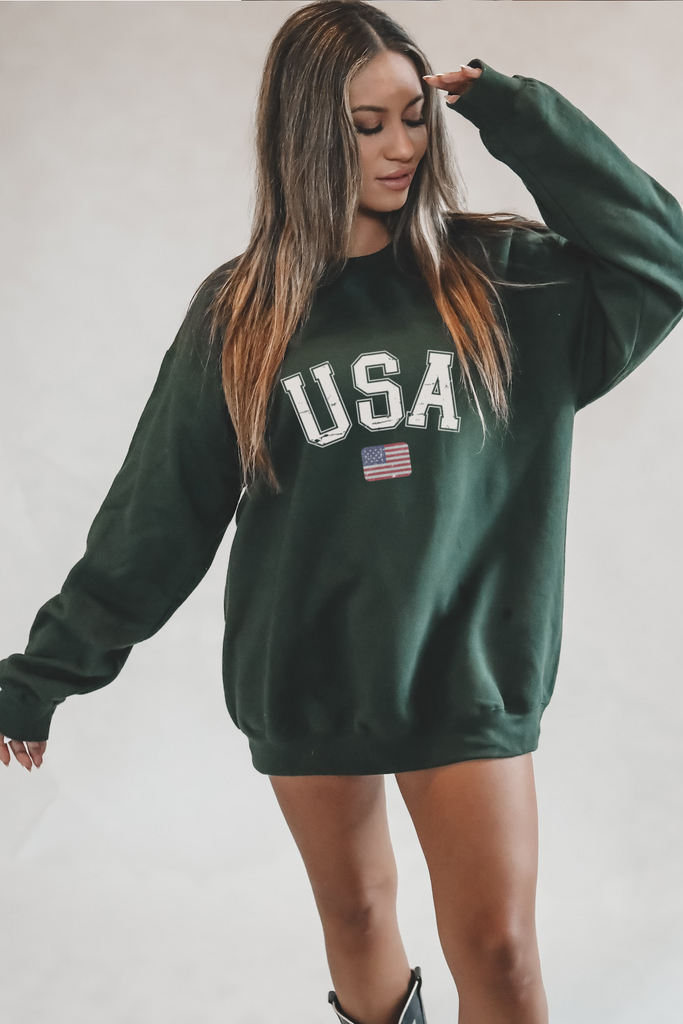 Person wearing a green sweatshirt with 'USA' text and an American flag, standing against a plain wall.