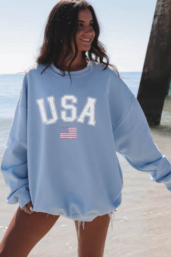 Woman wearing a blue 'USA' sweatshirt with an American flag on a beach.