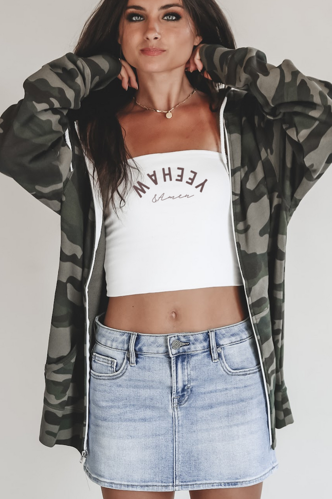 Woman wearing a camouflage jacket, white crop top, denim skirt, and cowboy boots on a plain background